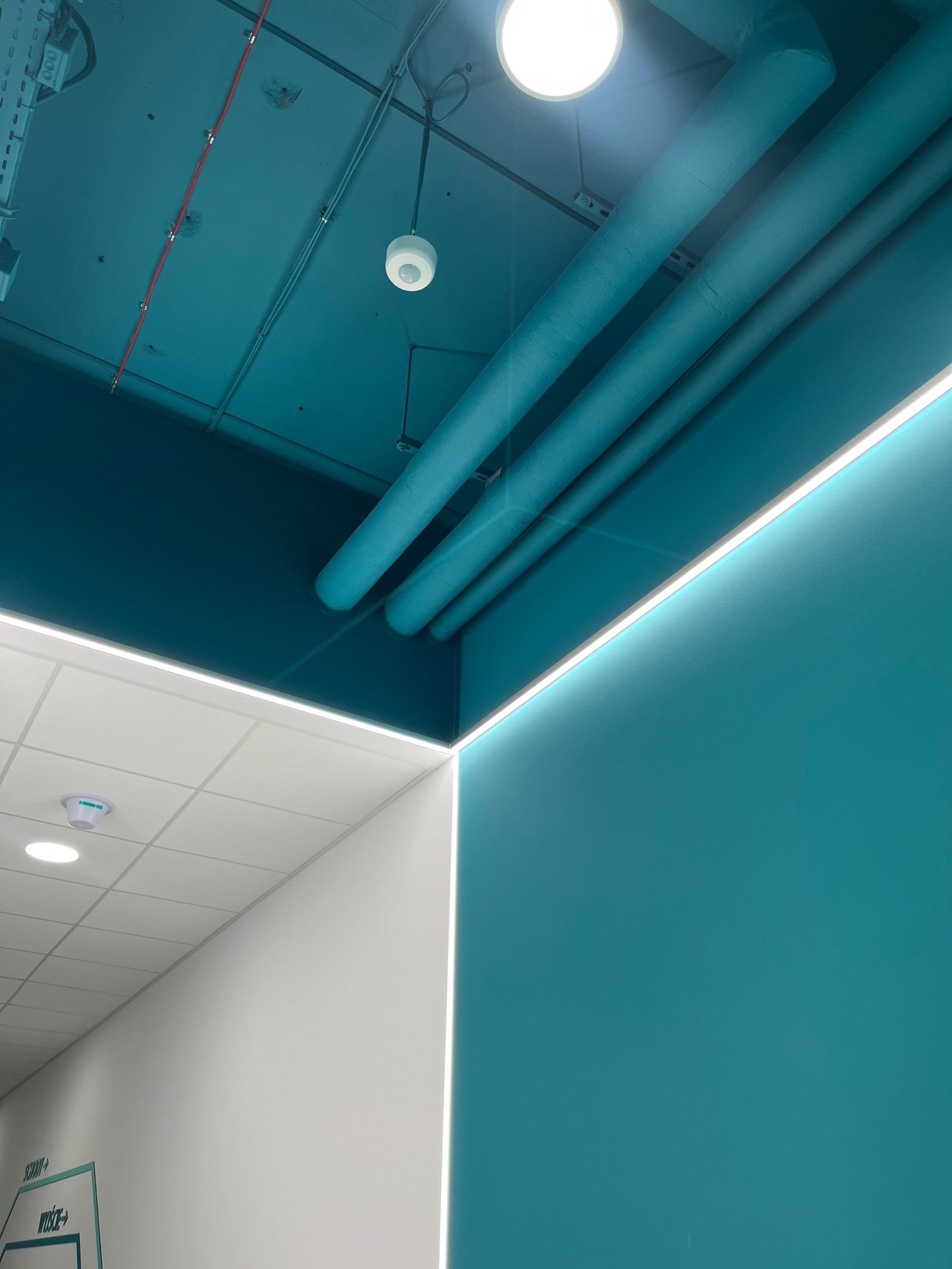 Narożnik: turkusowa ściana i strop z odsłoniętymi instalacjami obrysowane linią światła LED, obok biały korytarz z oznakowaniem