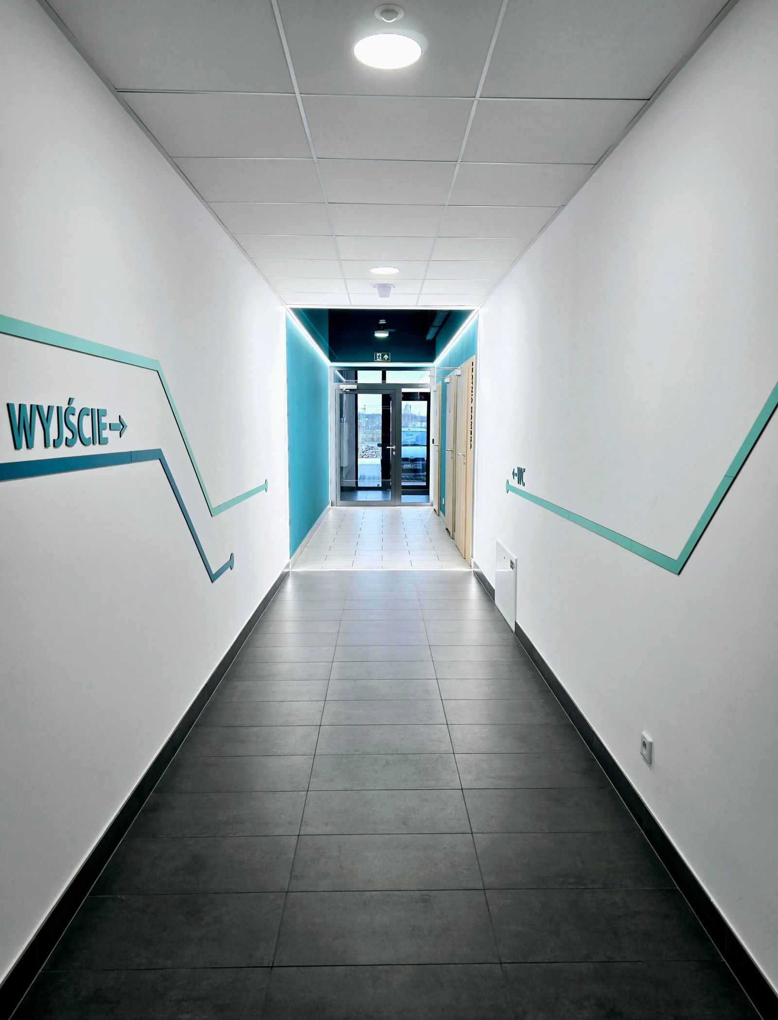 Korytarz z systemem informacji wizualnej: turkusowe linie na białych ścianach prowadzą do wyjścia i WC