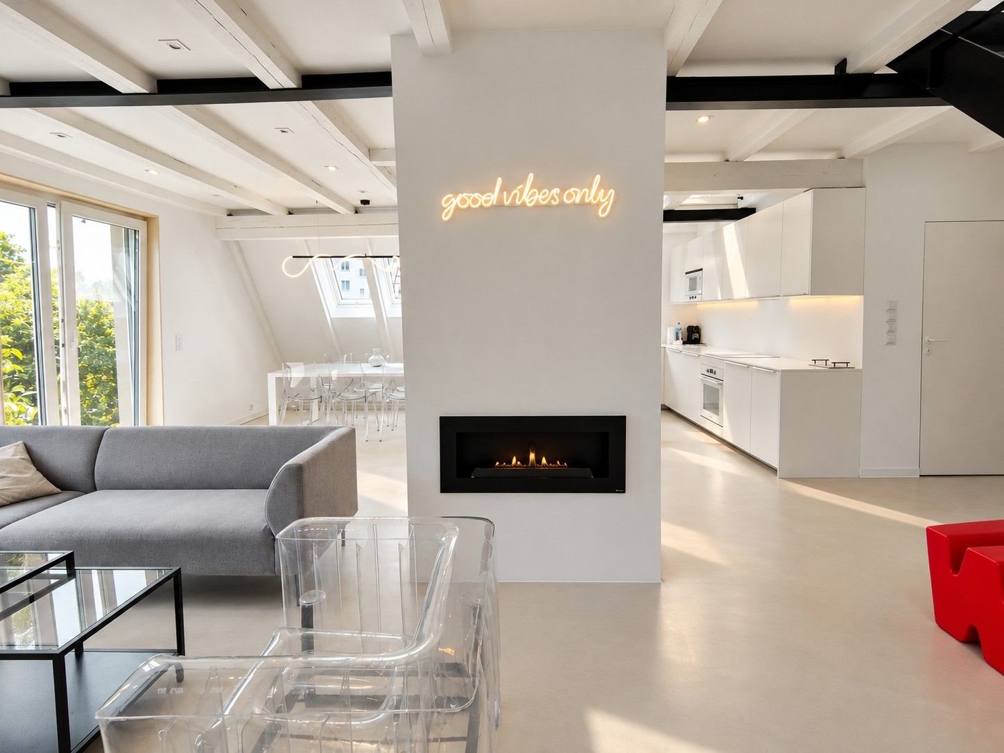 Salon z kominkiem w białej ścianie i neonem, szara sofa, przezroczysty fotel, w głębi jadalnia pod oknami dachowymi i biała kuchnia